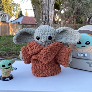 May include: A crocheted plush toy of a character from the Star Wars franchise, wearing a brown hooded jacket. The toy is sitting on a white surface next to a smaller plush toy of the same character.