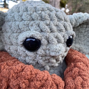 May include: A crocheted plush toy resembling a character from the Star Wars franchise. The toy has large, black eyes and a gray, textured body. The toy is wearing a brown, fuzzy scarf.