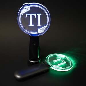 May include: Two illuminated LED light sticks with the letters "TI" inside a circle. The circle is outlined with a blue glow and has leaf-like designs. One stick is upright, the other is lying down, both emitting a green light.