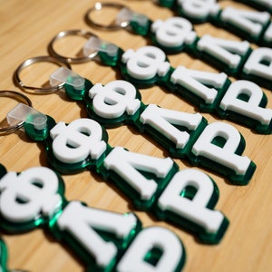May include: A set of green and white keychains with the letters "PHI" on them. The keychains are made of plastic and have a shiny finish.