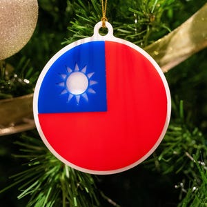 May include: A round Christmas ornament with the flag of Taiwan. The ornament is red, white, and blue with a blue sun in the upper left corner.