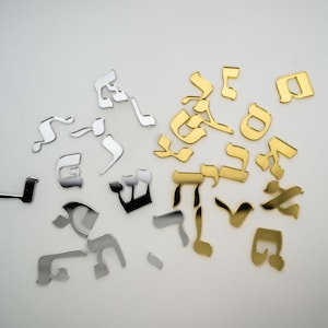 May include: Scattered across a white surface are metallic Hebrew letters in silver, gold, and black. The letters vary in size and style, with some appearing more stylized than others. The arrangement is random, suggesting a decorative or artistic purpose.