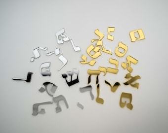 May include: Scattered across a white surface are metallic Hebrew letters in silver, gold, and black. The letters vary in size and style, with some appearing more stylized than others. The arrangement is random, suggesting a decorative or artistic purpose.