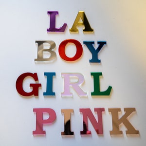 May include: Colorful acrylic letters spelling out the words "LA BOY GIRL PINK". The letters are arranged in a pyramid shape, with the largest letters at the top and the smallest letters at the bottom.