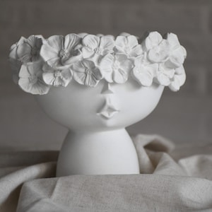 Puede incluir: Maceta de cerámica blanca con forma de cara y una corona de flores. La maceta tiene una base redonda y una parte superior ancha y abierta.