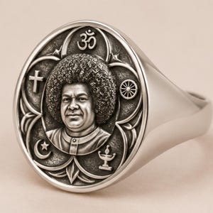 Op de afbeelding: Een zilverkleurige zegelring met een rond vlak voorzien van een reliëfportret van een persoon met krullend haar. Het portret is omringd door religieuze symbolen, waaronder een kruis, een Om, een ster met halve maan, een wiel en een lamp.