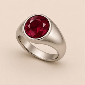 May include: A polished silver-tone signet ring featuring a large, round, faceted deep red gemstone set securely in the centre. The band is smooth and solid, with a rounded profile that tapers slightly towards the bottom of the ring's shank.