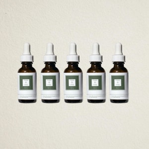 Könnte beinhalten: Fünf bernsteinfarbene Glasflaschen mit weißen Tropfern und Etiketten mit der Aufschrift "TAYA & COBA". Die Flaschen sind in einer Reihe auf einem hellbeigen Hintergrund angeordnet.