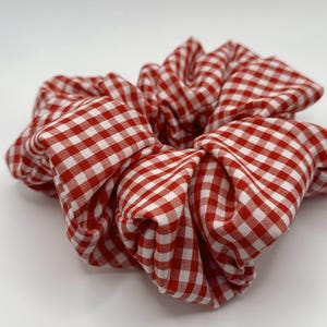 May include: A large red and white gingham scrunchie. The fabric is gathered and knotted, creating a voluminous hair accessory. The classic check pattern is evenly spaced and the scrunchie is set against a white background.