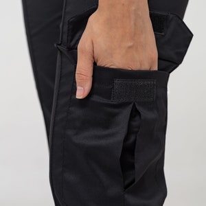 May include: Black cargo pants with a large flap pocket on the right leg. The pocket has a velcro closure.