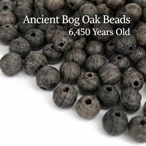 May include: A collection of ancient bog oak beads, approximately 6,450 years old. The beads are a dark gray color with a natural wood grain texture. Each bead is round with a drilled hole. The text "Ancient Bog Oak Beads 6,450 Years Old" is visible.