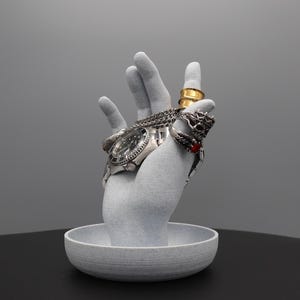 May include: A grey, textured, hand-shaped ring and watch holder with a small, grey, textured dish at the base. The holder has a silver watch and several rings on the fingers.