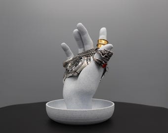May include: A grey, textured, hand-shaped ring and watch holder with a small, grey, textured dish at the base. The holder has a silver watch and several rings on the fingers.