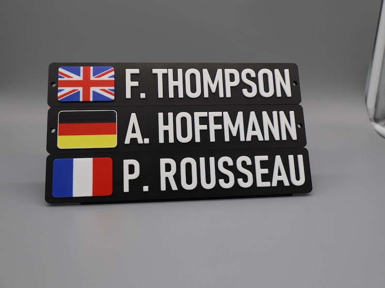 Three signs on a stand for display purposes, with a grey background. Each sign has a country flag on the left side, followed by uppercase text. The text consists of the first letter of the first name, a dot and the full surname.