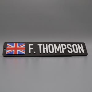 A single sign on a stand for display purposes, in with a grey background. The sign has the UK flag on the left side, followed by the text "F. THOMPSON".