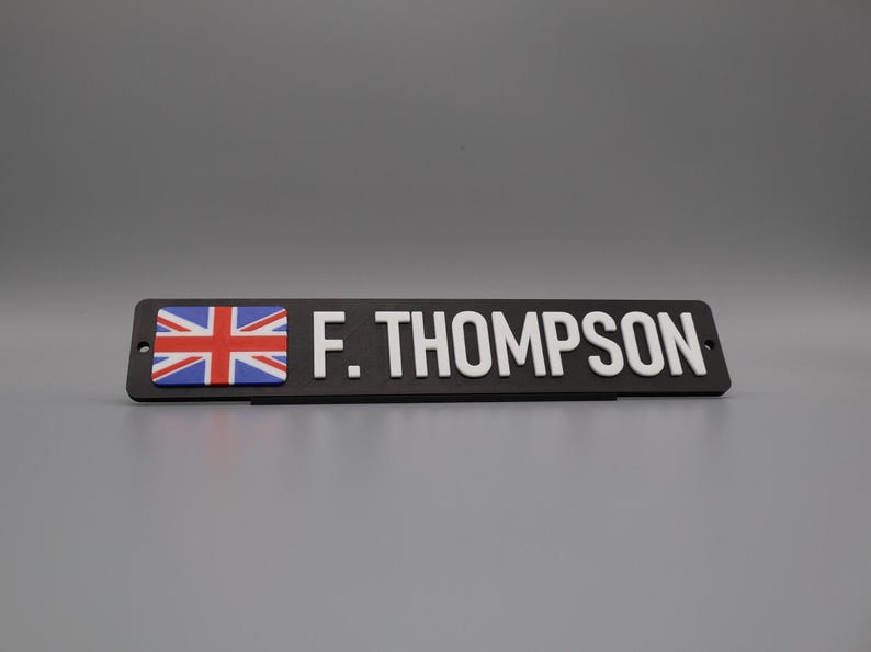 A single sign on a stand for display purposes, in with a grey background. The sign has the UK flag on the left side, followed by the text "F. THOMPSON".