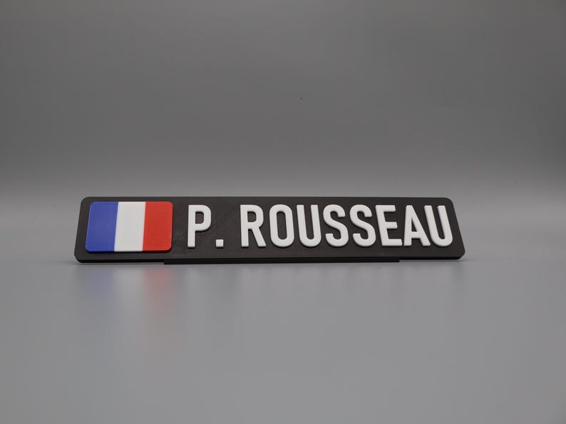 A single sign on a stand for display purposes, in with a grey background. The sign has the flag of france on the left side, followed by the text "P. ROUSSEAU".