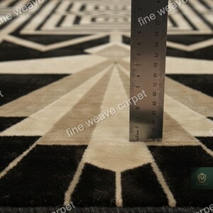Pu&ograve; includere: Tappeto con motivo geometrico in nero, beige e grigio. Il tappeto presenta un design simmetrico con linee radiali e forme geometriche. Il testo "fine weave carpet" &egrave; visibile. Un righello &egrave; posto sul tappeto.