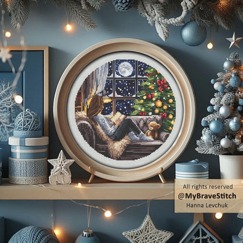 Puede incluir: Una obra de arte de punto de cruz en un marco de madera redondo representa a una persona leyendo junto a una ventana con un &aacute;rbol de Navidad y un oso de peluche. La escena est&aacute; ambientada contra un cielo estrellado. La obra se exhibe en una estanter&iacute;a con otras decoraciones navide&ntilde;as.