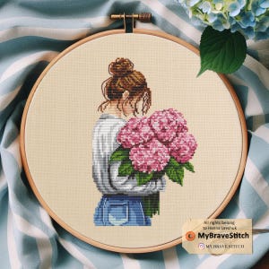 Puede incluir: Patrón de punto de cruz de una mujer con cabello castaño sosteniendo un ramo de hortensias rosas. El patrón está en un aro de bordado de madera. El texto "MyBraveStitch" está en la esquina inferior derecha de la imagen.