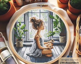 May include: A cross-stitch embroidery hoop featuring a woman meditating in front of a window, surrounded by potted plants. The design is in shades of gray, blue, and brown, with a wooden hoop. The text "All rights reserved @MyBraveStitch Hanna Levchuk" is visible.