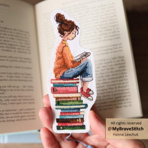 Peut inclure: Un marque-page en point de croix représentant une personne avec un chignon brun, portant un pull orange et un jean bleu, assise sur une pile de livres colorés en lisant. Le marque-page a une bordure en tissu blanc et un pompon bleu en bas.