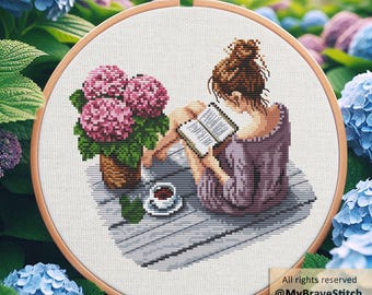 May include: Cross-stitch embroidery in a wooden hoop depicts a person reading a book outdoors. A cup of coffee and a pot of pink hydrangeas are nearby. The text "All rights reserved @MyBraveStitch Hanna Levchuk" is in the lower right corner.