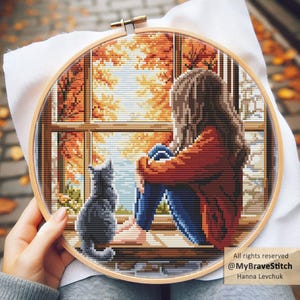 Puede incluir: Un bordado de punto de cruz en un aro de madera representa a una persona mirando por una ventana el follaje otoñal. Un gato gris está sentado en el alféizar. La escena está bordada con tonos de naranja, marrón, azul y gris. El texto "@MyBraveStitch" y "Hanna Levchuk" son visibles.