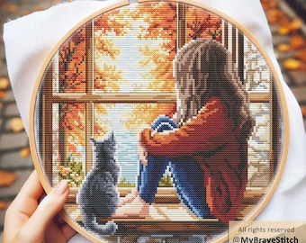 May include: A cross-stitch embroidery in a wooden hoop depicts a person looking out a window at autumn foliage. A gray cat sits on the windowsill. The scene is stitched with shades of orange, brown, blue, and gray. The text "@MyBraveStitch" and "Hanna Levchuk" are visible.