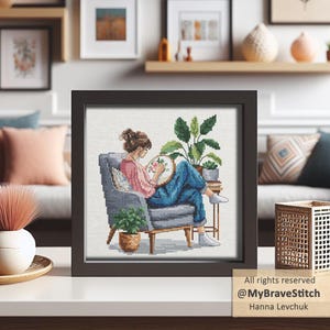 May include: Framed cross-stitch art depicts a person seated in a gray armchair, engrossed in embroidery. The artwork features a person wearing a pink shirt and blue jeans, surrounded by potted plants. The frame is dark brown, and the image is displayed on a white surface.
