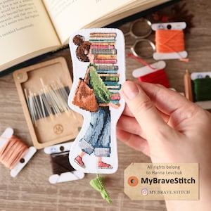 Peut inclure: Marque-page en point de croix représentant une femme portant une pile de livres, vêtue d'une chemise verte, d'un jean bleu et de chaussures rouges. Elle porte un sac marron. Autour, des fournitures de couture.