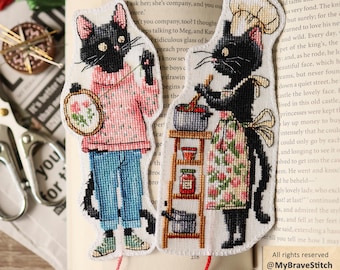 Könnte beinhalten: Zwei Kreuzstich-Lesezeichen mit schwarzen Katzen. Die linke Katze trägt einen rosa Pullover und blaue Jeans und hält einen Stickrahmen. Die rechte Katze trägt eine Kochmütze und eine geblümte Schürze und kocht an einem Regal mit Gläsern.