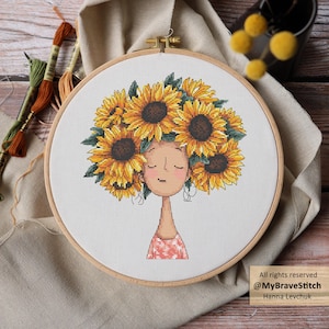 Puede incluir: Bordado de punto de cruz de una mujer con una cabeza de girasoles, en un aro de madera. El diseño presenta una cara con los ojos cerrados y una parte superior rosa estampada. El texto "All rights reserved @MyBraveStitch Hanna Levchuk" es visible.