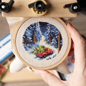 May include: A circular cross-stitch embroidery project mounted in a wooden hoop, depicting a red pickup truck carrying a green Christmas tree through a snowy forest landscape with tall dark blue and brown trees under a winter sky.