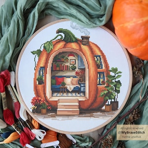May include: A cross-stitch embroidery in a round wooden hoop depicting a large orange pumpkin house with a cozy interior, including a bookshelf, blue furniture, and a chimney with smoke, surrounded by green fabric and embroidery floss.