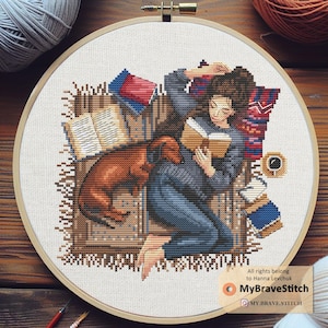 Puede incluir: Bordado de punto de cruz en un aro de madera que representa a una persona leyendo con un perro salchicha, rodeada de libros y una taza de café. El diseño presenta una escena acogedora con colores cálidos y detalles intrincados.