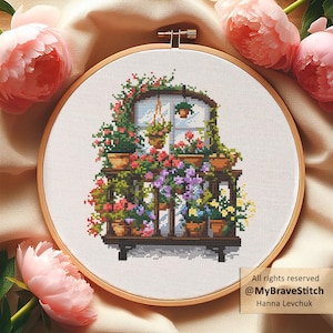 Op de afbeelding: Kruissteekborduurwerk van een bloembak vol kleurrijke bloemen in potten. Het ontwerp heeft een houten frame, een raam met uitzicht en een verscheidenheid aan bloemen in tinten rood, roze, paars en geel. De borduurwerk is in een houten hoepel.