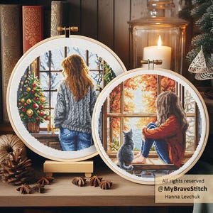 Puede incluir: Dos bastidores redondos de punto de cruz con escenas de una persona mirando por ventanas. Uno muestra una escena invernal con un árbol de Navidad, el otro una escena otoñal con un gato. Los bastidores se exhiben sobre una superficie de madera.
