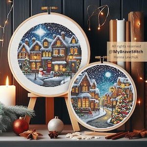 Op de afbeelding: Twee ingelijste kruissteekkunstwerken tonen een besneeuwd dorpsgezicht met verlichte huizen, een sneeuwpop en een kerstboom. Het kunstwerk staat tegen een donkere achtergrond met decoratieve elementen, waaronder een kaars, ornamenten en kaneelstokjes. Tekst: All rights reserved @MyBraveStitch Hanna Levchuk.