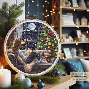 Puede incluir: Un bastidor de bordado de punto de cruz con una escena navide&ntilde;a acogedora. El dise&ntilde;o representa a una persona leyendo junto a una ventana con vistas a un &aacute;rbol de Navidad decorado y una luna llena. Un osito de peluche est&aacute; cerca. El bastidor se encuentra sobre un fondo c&aacute;lido.