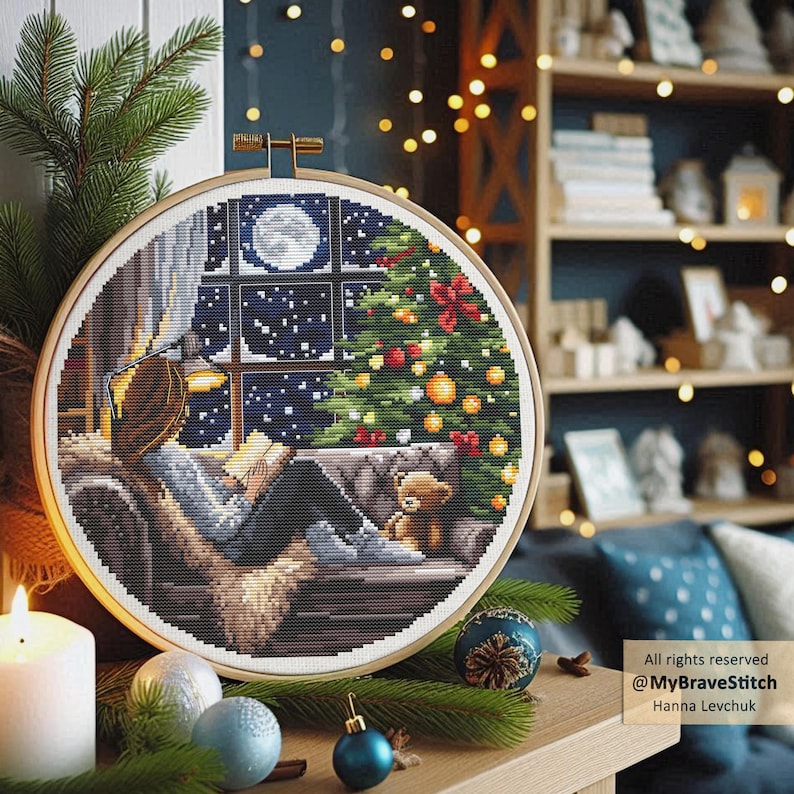 Puede incluir: Un bastidor de bordado de punto de cruz con una escena navide&ntilde;a acogedora. El dise&ntilde;o representa a una persona leyendo junto a una ventana con vistas a un &aacute;rbol de Navidad decorado y una luna llena. Un osito de peluche est&aacute; cerca. El bastidor se encuentra sobre un fondo c&aacute;lido.