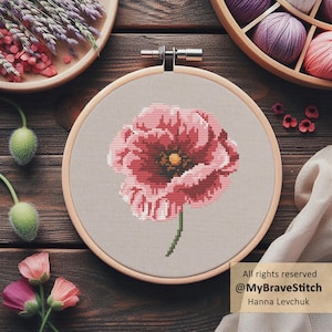 Può includere: Ricamo a punto croce di un fiore di papavero rosa in un telaio di legno. Il fiore ha un centro giallo e uno stelo verde. Lo sfondo è un tessuto grigio chiaro. L'immagine include il testo "All rights reserved @MyBraveStitch Hanna Levchuk."