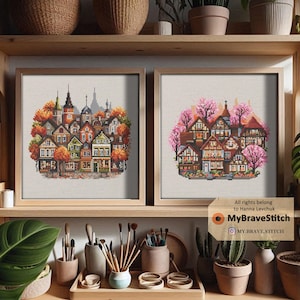 May include: Two framed cross-stitch pictures showcasing detailed European townscapes. One has autumnal colours, the other pink blossom. The artwork is displayed on a wooden shelf with art supplies and decorative items.