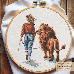 May include: A cross-stitch embroidery in a wooden hoop shows a person walking a lion on a lead. The person wears a rust-coloured jumper and blue jeans. The lion is brown with a large mane. The text "All rights reserved @MyBraveStitch Hanna Levchuk" is in the lower right.