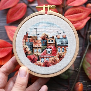 May include: A circular wooden embroidery hoop containing a cross-stitch design of a townscape with houses featuring red, orange, and blue tones. The scene includes rooftops, windows, and aerials against a light beige fabric background.