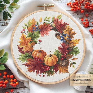 May include: Cross-stitch embroidery in a wooden hoop features a fall wreath design. The wreath includes orange pumpkins, red berries, and colorful autumn leaves in shades of orange, yellow, and red. The text "All rights reserved @MyBraveStitch Hanna Levchuk" is visible.