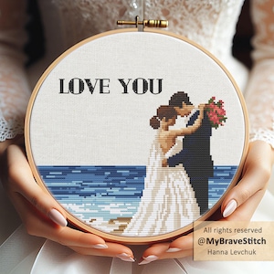 Puede incluir: Un bastidor de bordado de punto de cruz con una escena de boda con una pareja abrazándose, sobre un fondo de océano azul. Las palabras "LOVE YOU" están bordadas en la parte superior. El bastidor es sostenido por una persona.