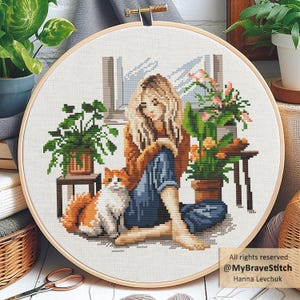 Op de afbeelding: Kruissteekborduurwerk in een houten hoepel toont een persoon met lang blond haar, zittend met een kat, omringd door potplanten. De scène speelt zich af bij een raam. Het borduurwerk is in tinten bruin, blauw, groen en oranje.