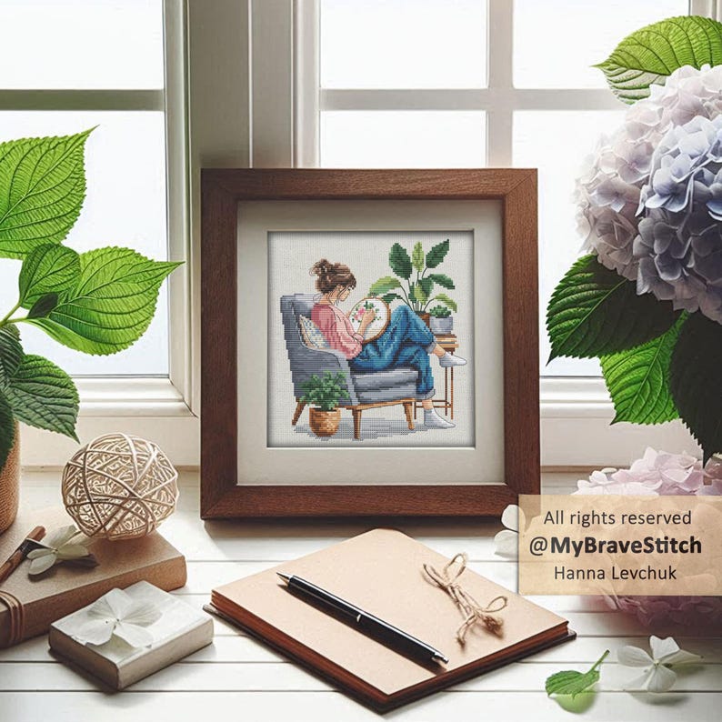 May include: A framed cross-stitch artwork depicts a person seated in a gray armchair, engrossed in embroidery. The artwork is set against a window with greenery and a bouquet of flowers. A notepad, pen, and decorative items are in the foreground. Text: All rights reserved @MyBraveStitch Hanna Levchuk.