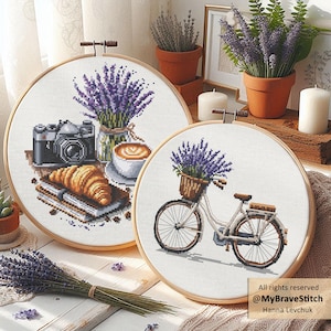Puede incluir: Dos bastidores de bordado de punto de cruz con temas de lavanda. Uno presenta una cámara, un cruasán y café. El otro representa una bicicleta con una cesta de lavanda. Ambos están enmarcados en aros de madera.